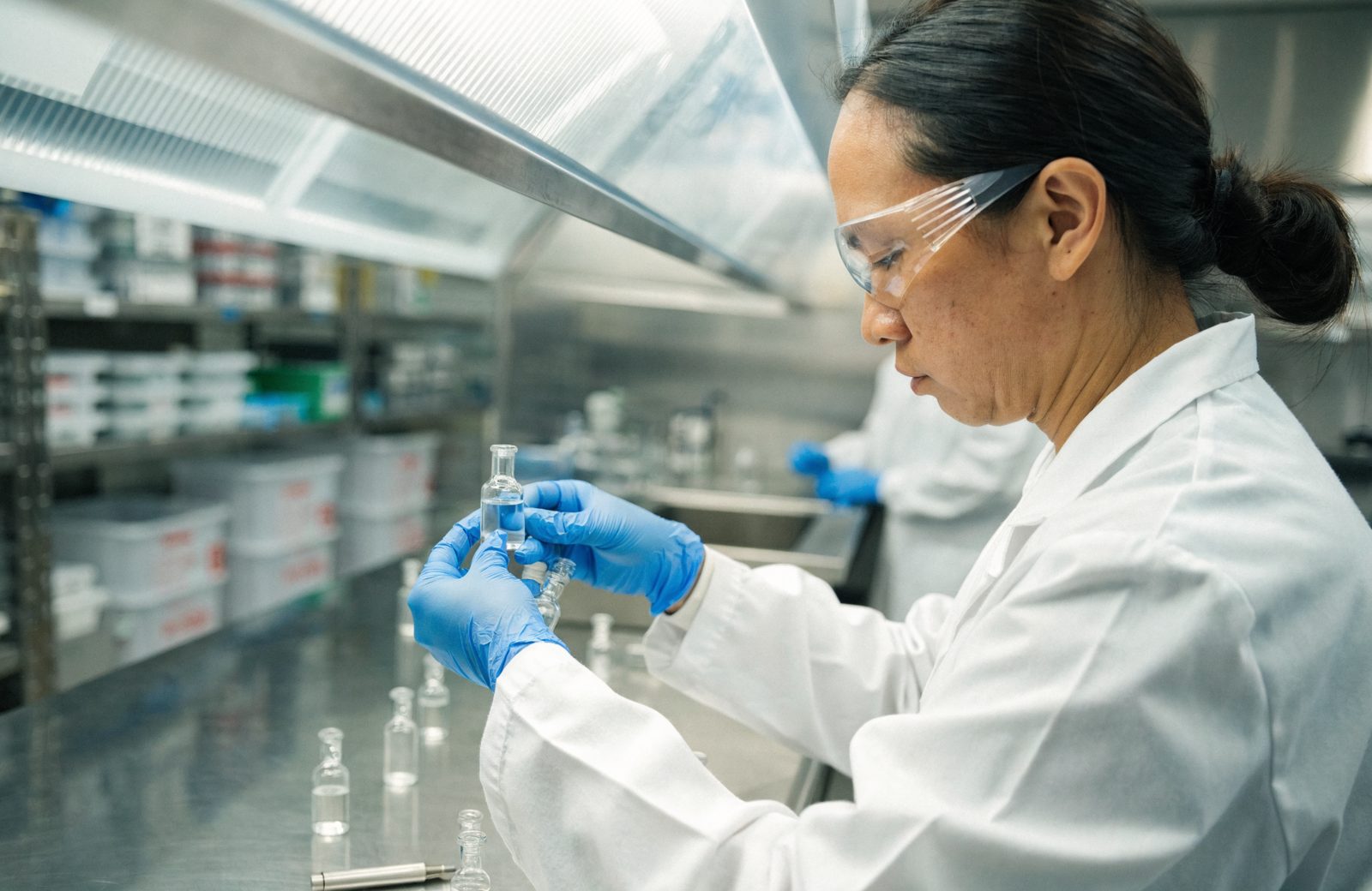 A pharmacy technician in a lab coat and gloves inspecting a vial in a compounding lab